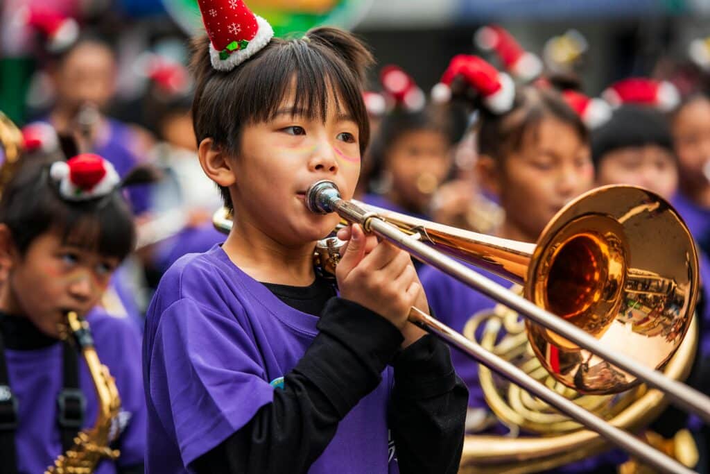 Child playing trombone in a festive parade, wearing a holiday hat, outdoors.