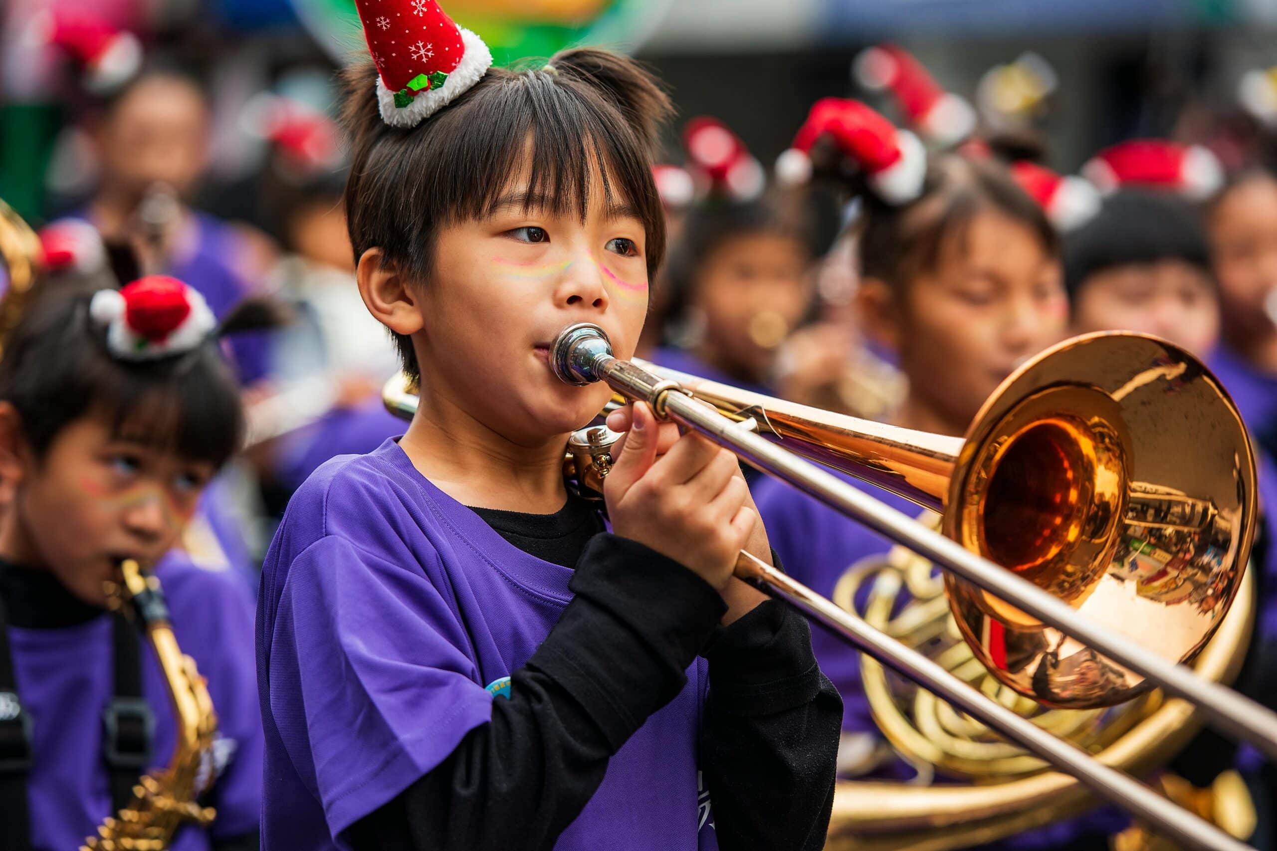 Child playing trombone in a festive parade, wearing a holiday hat, outdoors.