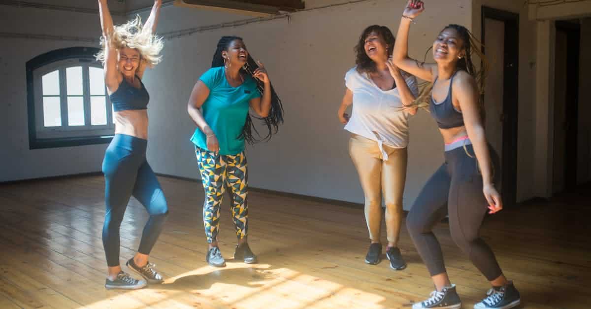 Four diverse women joyfully dancing together in a sunlit studio room.